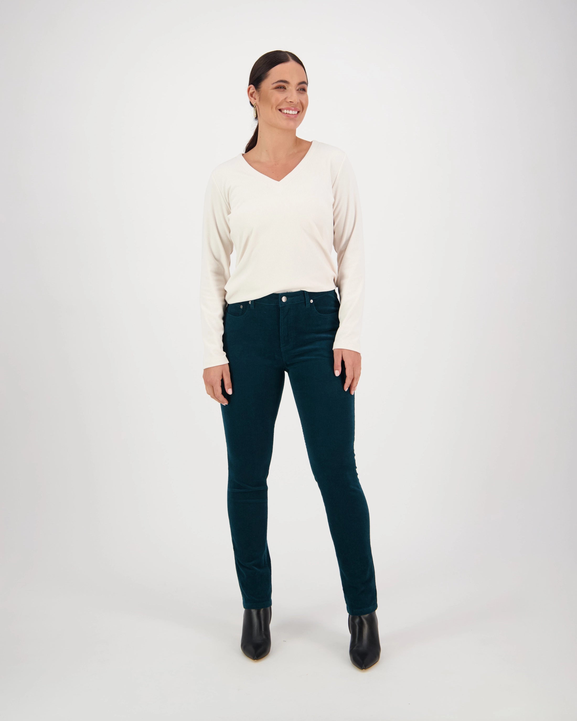 A woman smiles against a white background, wearing a cream long-sleeve V-neck sweater, black ankle boots, and Vassalli Park Cord Pant in Peacock.