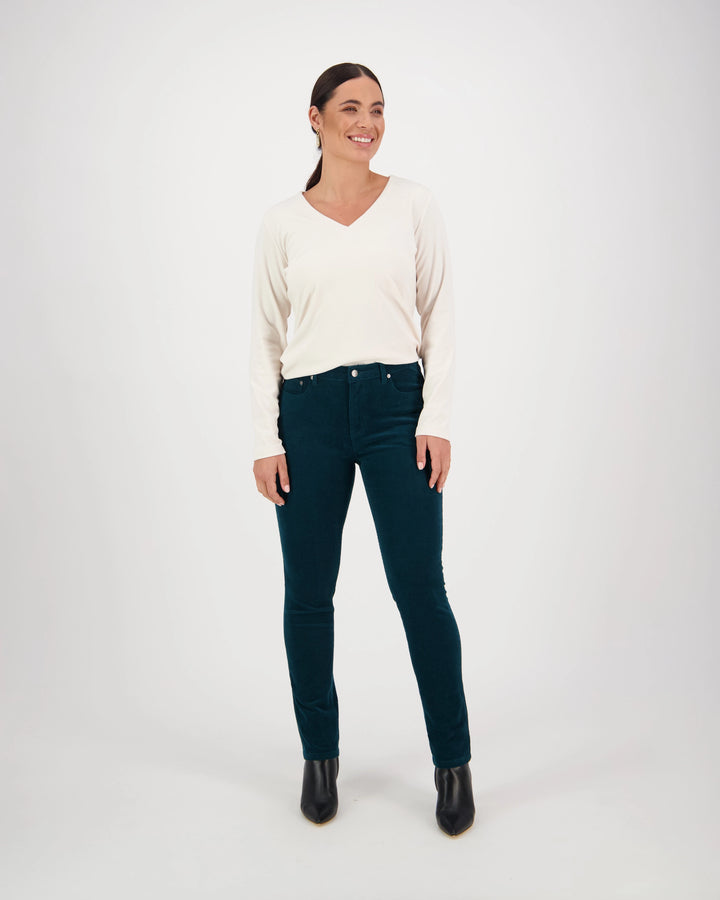 A woman smiles against a white background, wearing a cream long-sleeve V-neck sweater, black ankle boots, and Vassalli Park Cord Pant in Peacock.