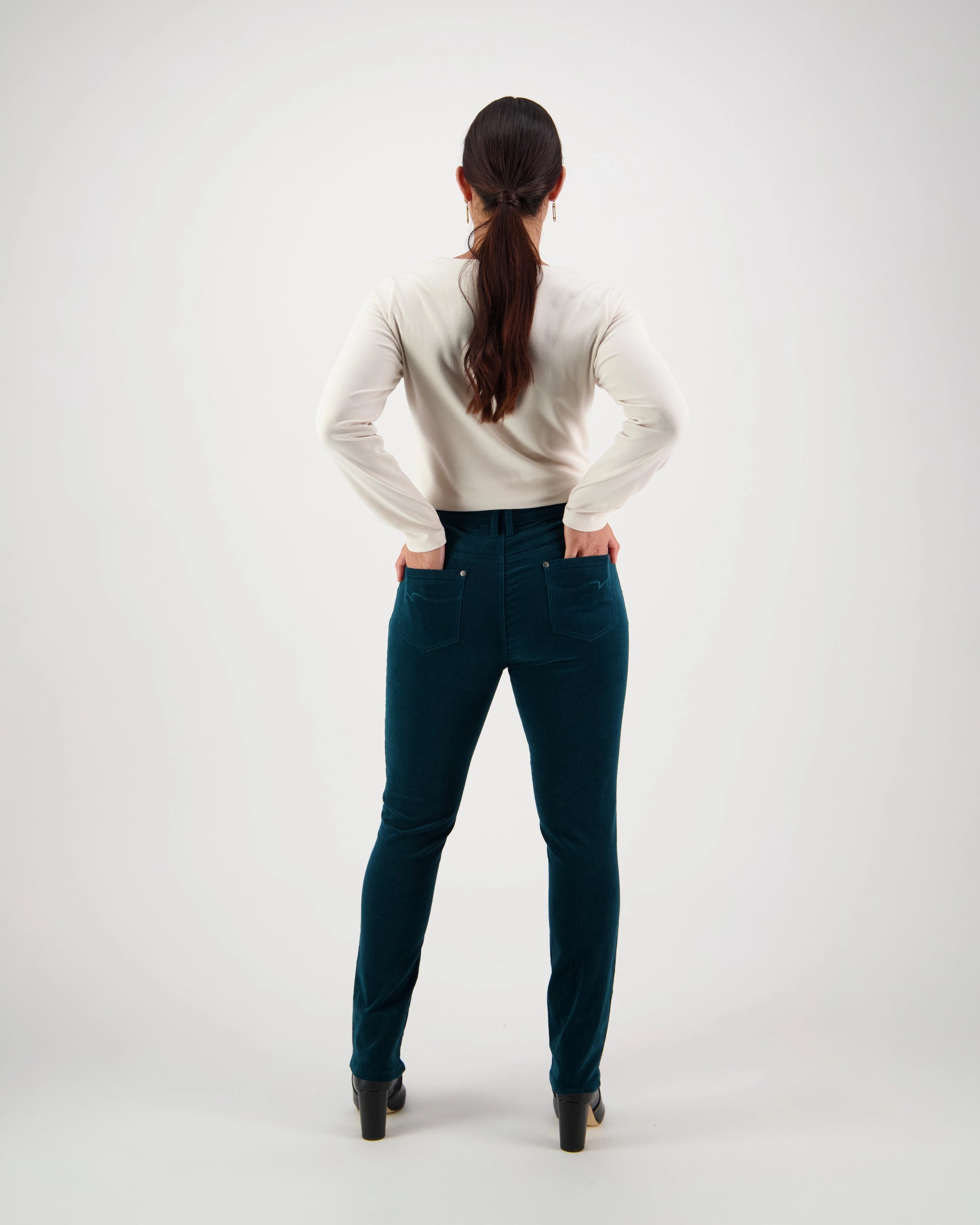 A woman with a ponytail stands facing away on a white background, wearing the Vassalli Park Cord Pant - Peacock, a teal pinwale cord narrow-leg pant, paired with a white long-sleeve shirt and black heels.