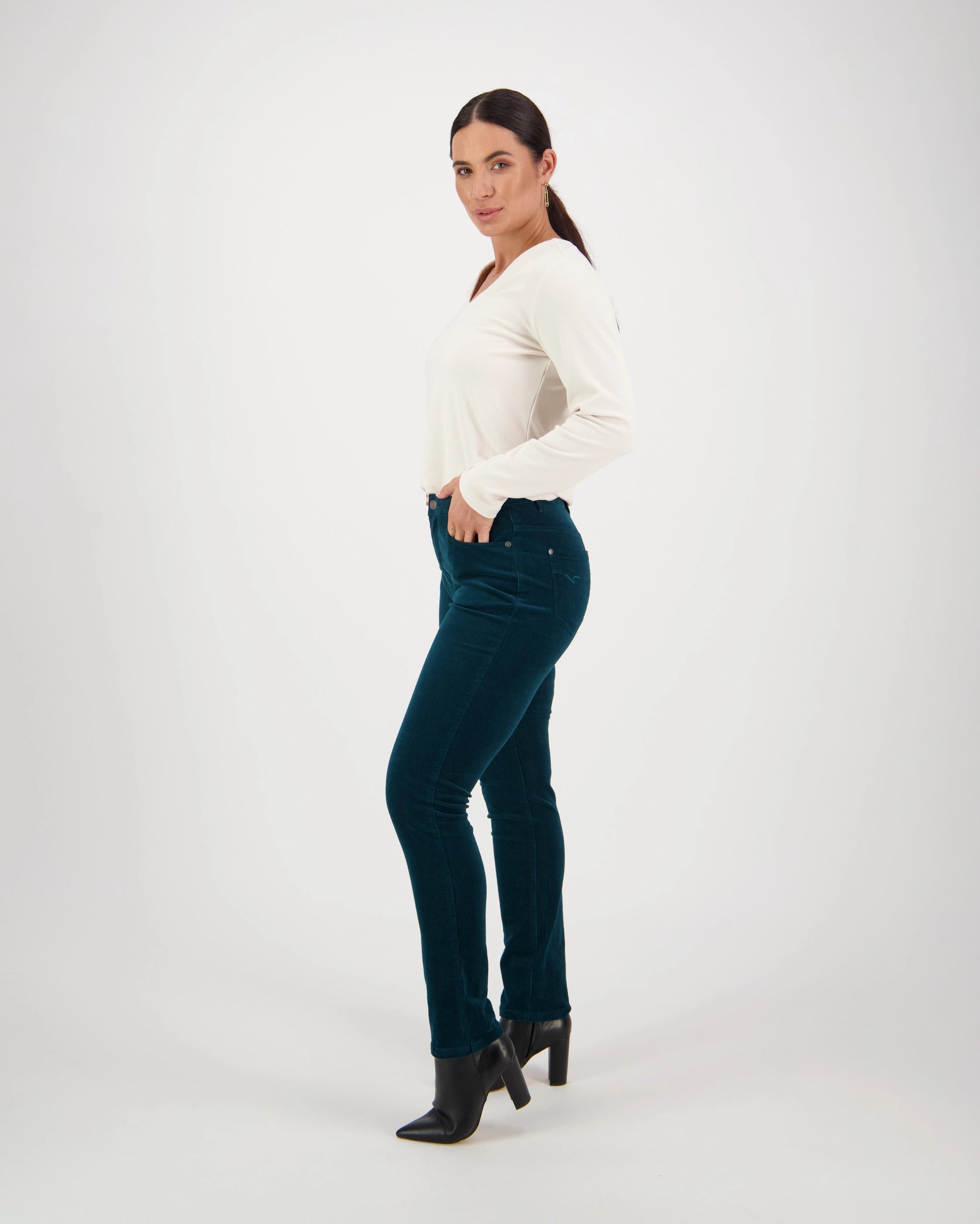 A woman poses confidently in a studio with a white background, wearing Vassalli’s Park Cord Pant in Peacock, a long-sleeve white top, and black heeled boots, with one hand in her pocket as she looks at the camera.