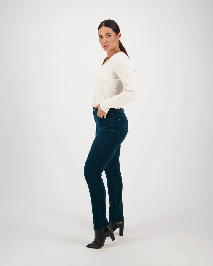 A woman poses confidently in a studio with a white background, wearing Vassalli’s Park Cord Pant in Peacock, a long-sleeve white top, and black heeled boots, with one hand in her pocket as she looks at the camera.