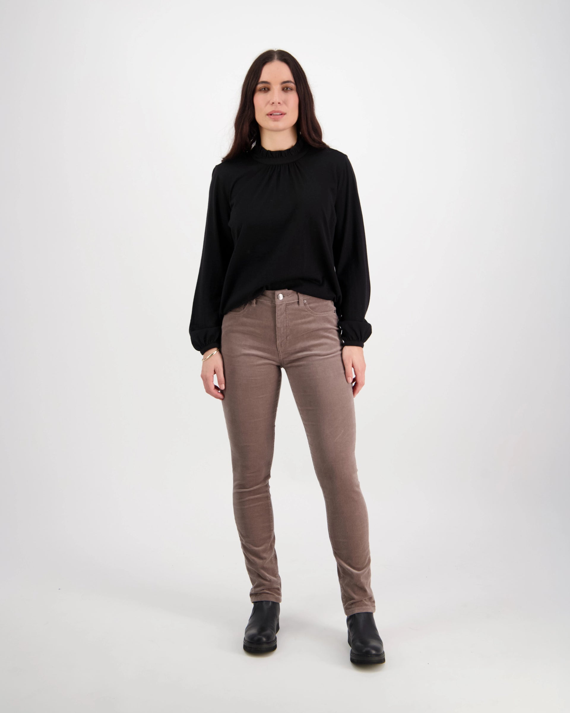 A woman with long dark hair faces the camera in a black long-sleeve top, black boots, and Vassalli’s Park Cord Pant in Taupe, against a plain white background.