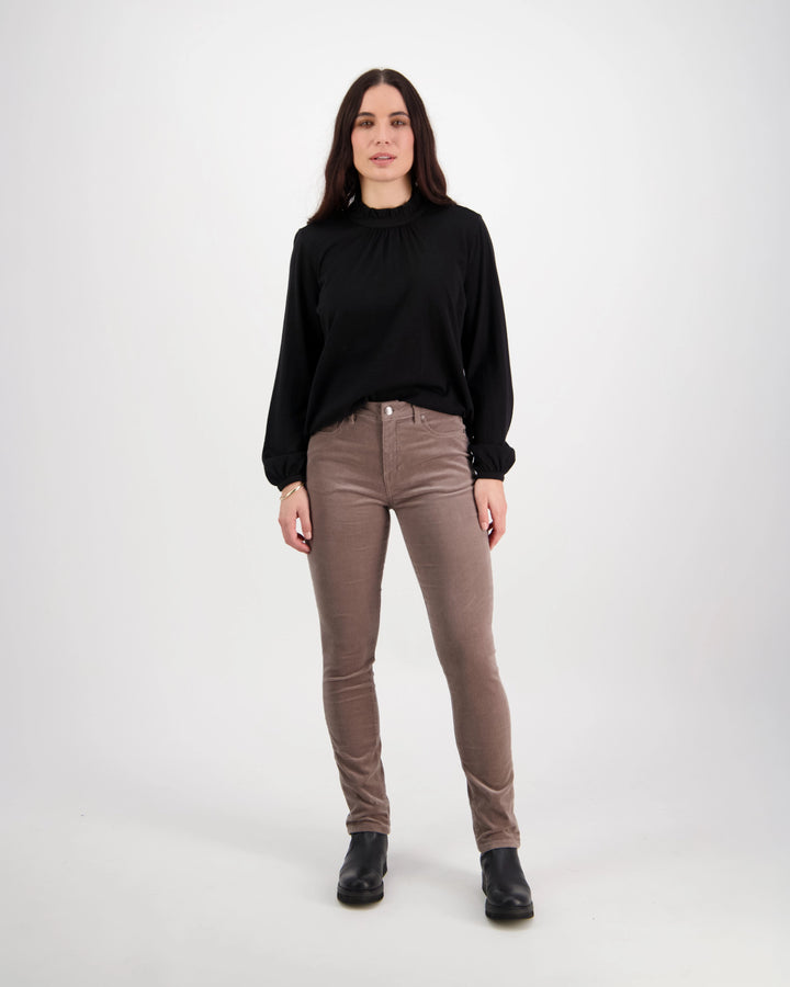 A woman with long dark hair faces the camera in a black long-sleeve top, black boots, and Vassalli’s Park Cord Pant in Taupe, against a plain white background.
