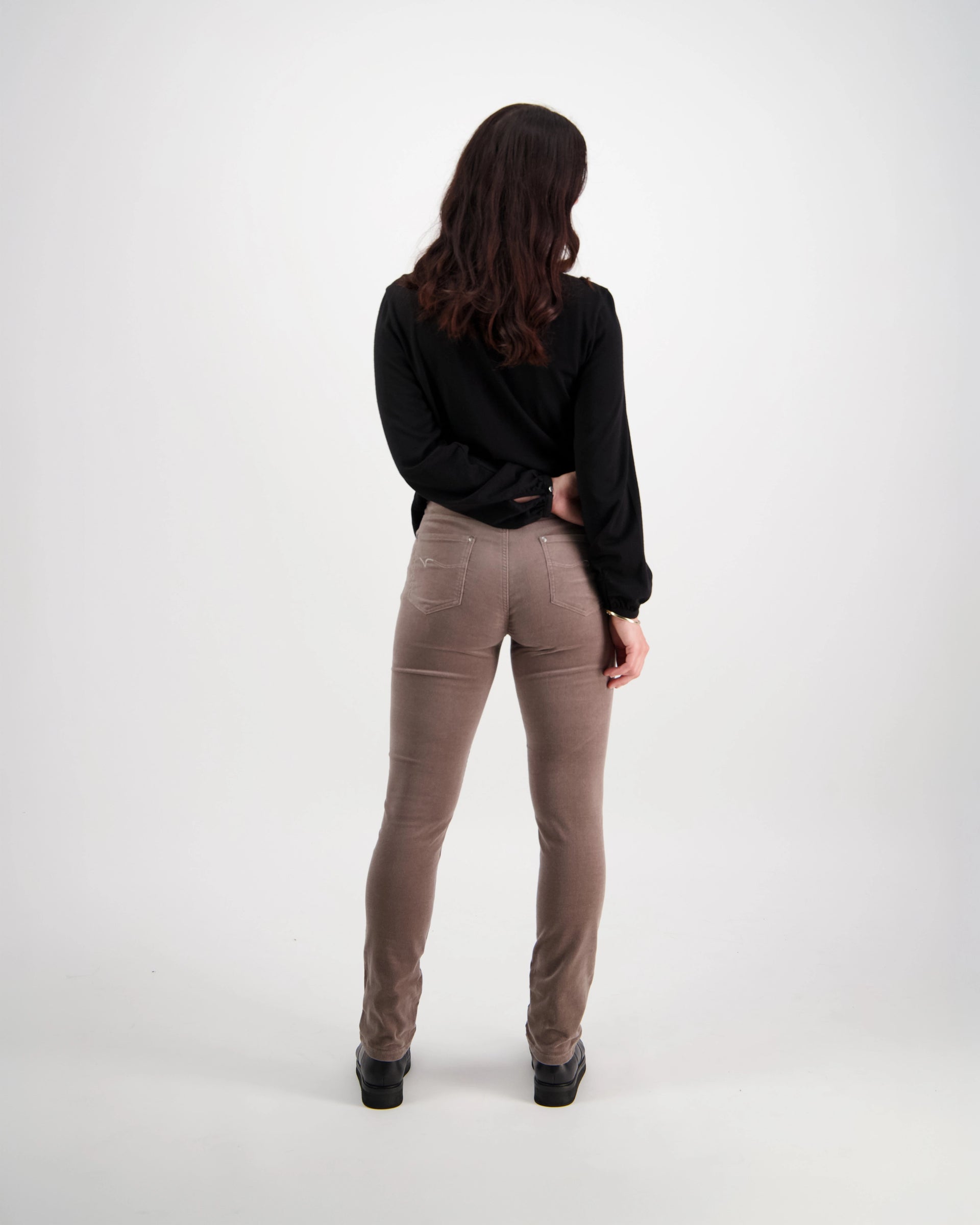A woman with long brown hair wears a black long-sleeve top and Vassalli’s Park Cord Pant in Taupe, standing with her back to the camera against a plain white background.