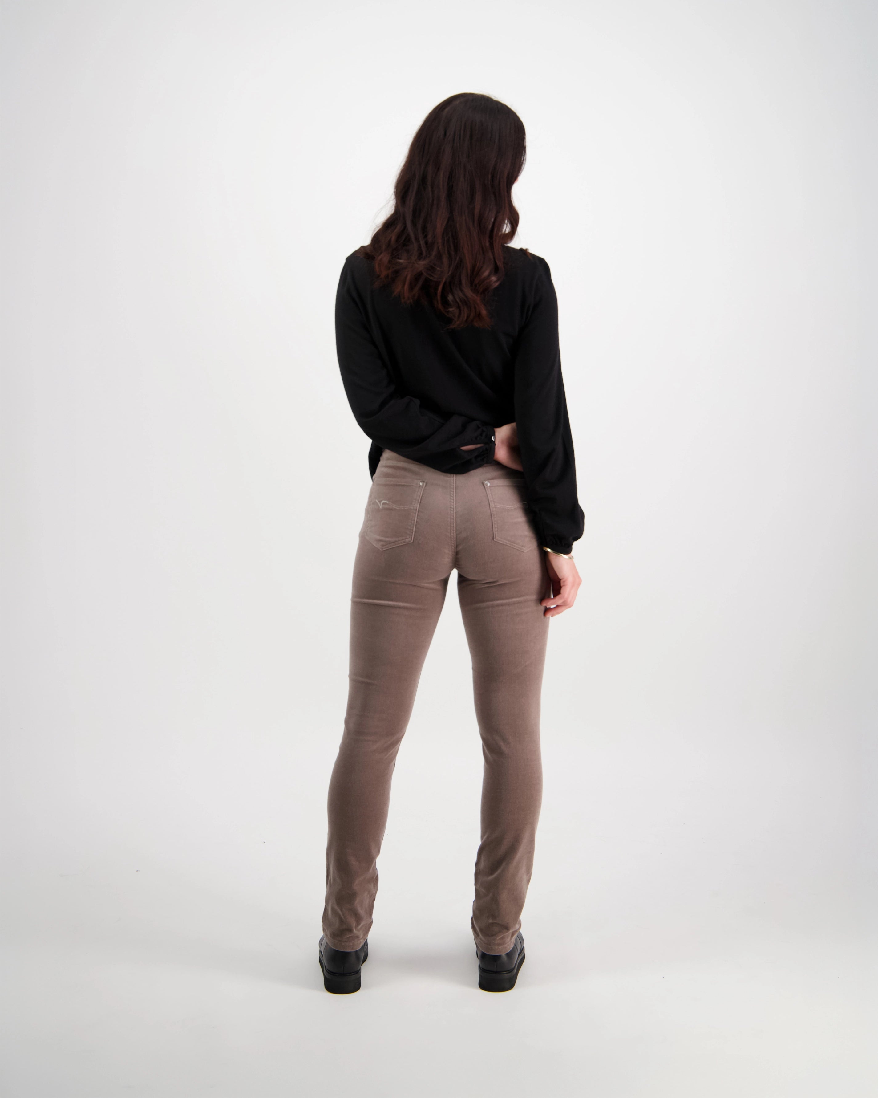 A woman with long brown hair wears a black long-sleeve top and Vassalli’s Park Cord Pant in Taupe, standing with her back to the camera against a plain white background.