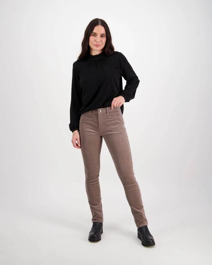 A woman with long dark hair stands against a plain white background, wearing a black long-sleeve top, Vassalli Park Cord Pant in taupe, and black ankle boots. She is smiling slightly.