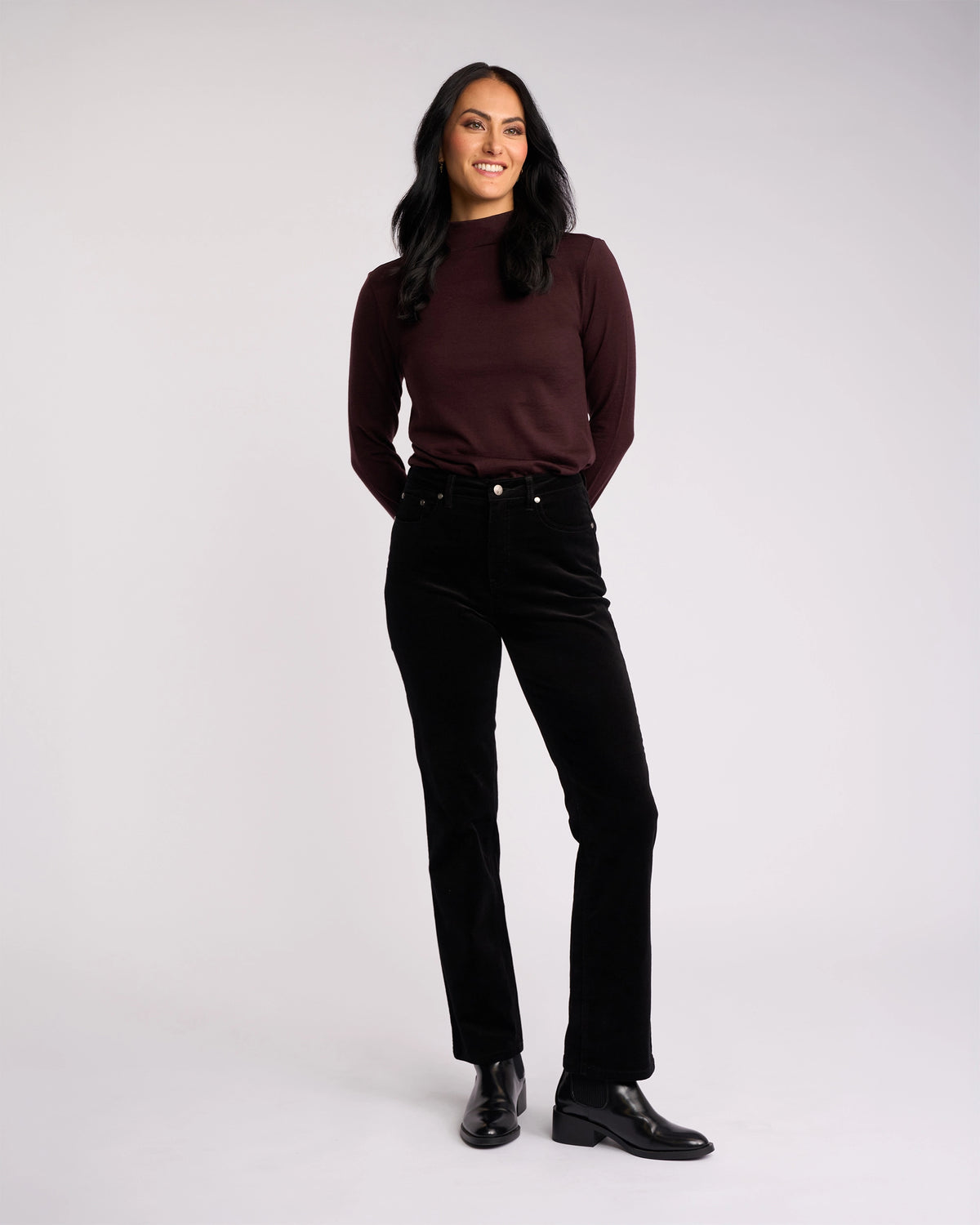A woman with long dark hair, wearing a maroon top and Vassalli’s Arch Cord Pant in black, pairs her look with black ankle boots while smiling against a light background—a great option for your winter wardrobe.