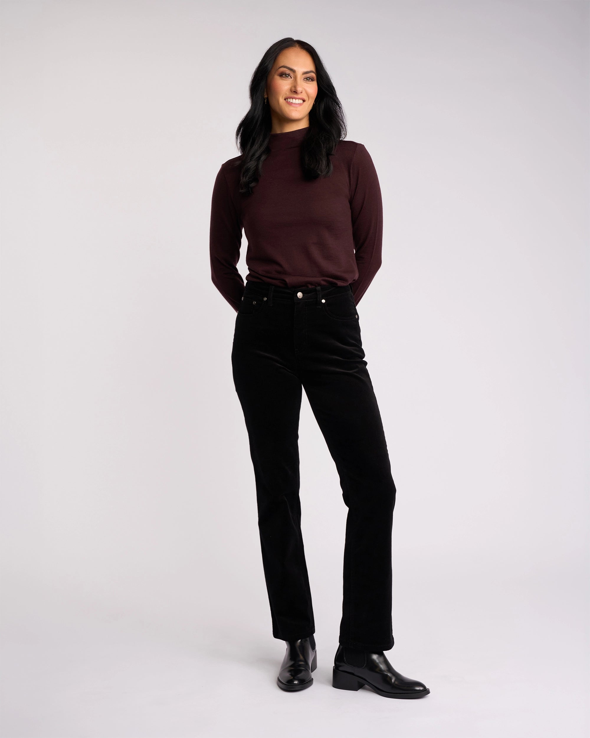A woman with long dark hair, wearing a maroon top and Vassalli’s Arch Cord Pant in black, pairs her look with black ankle boots while smiling against a light background—a great option for your winter wardrobe.