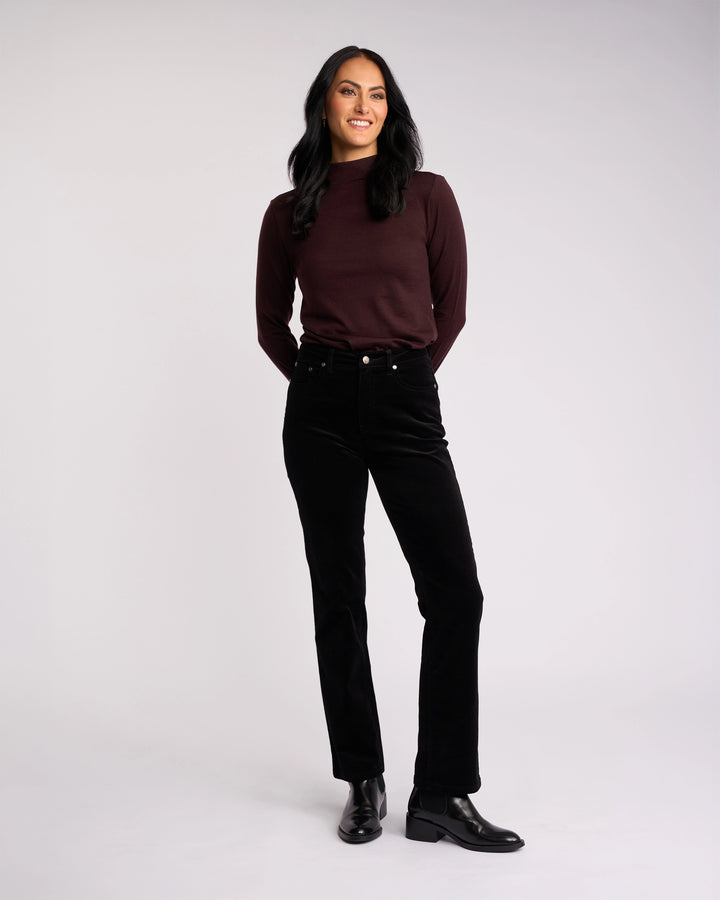 A woman with long dark hair, wearing a maroon top and Vassalli’s Arch Cord Pant in black, pairs her look with black ankle boots while smiling against a light background—a great option for your winter wardrobe.