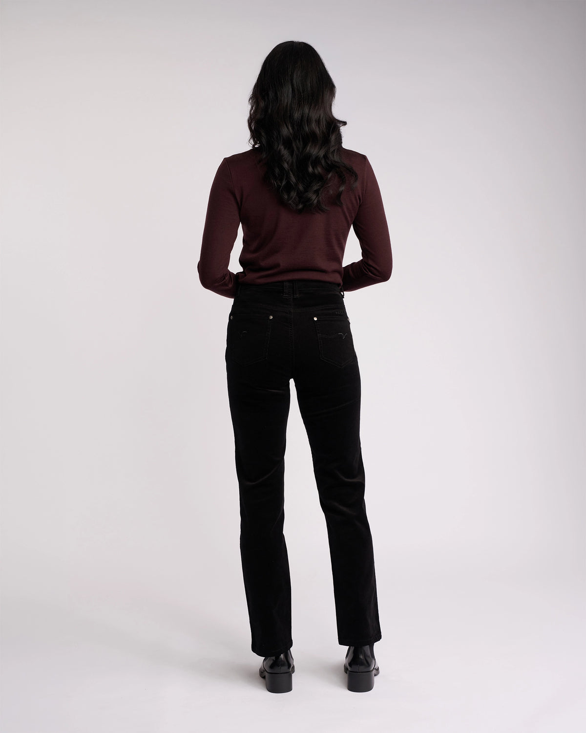 A person with long dark hair, wearing a maroon long-sleeve top and Vassalli Arch Cord Pant in Black, stands facing away against a plain light background—an easy winter style.