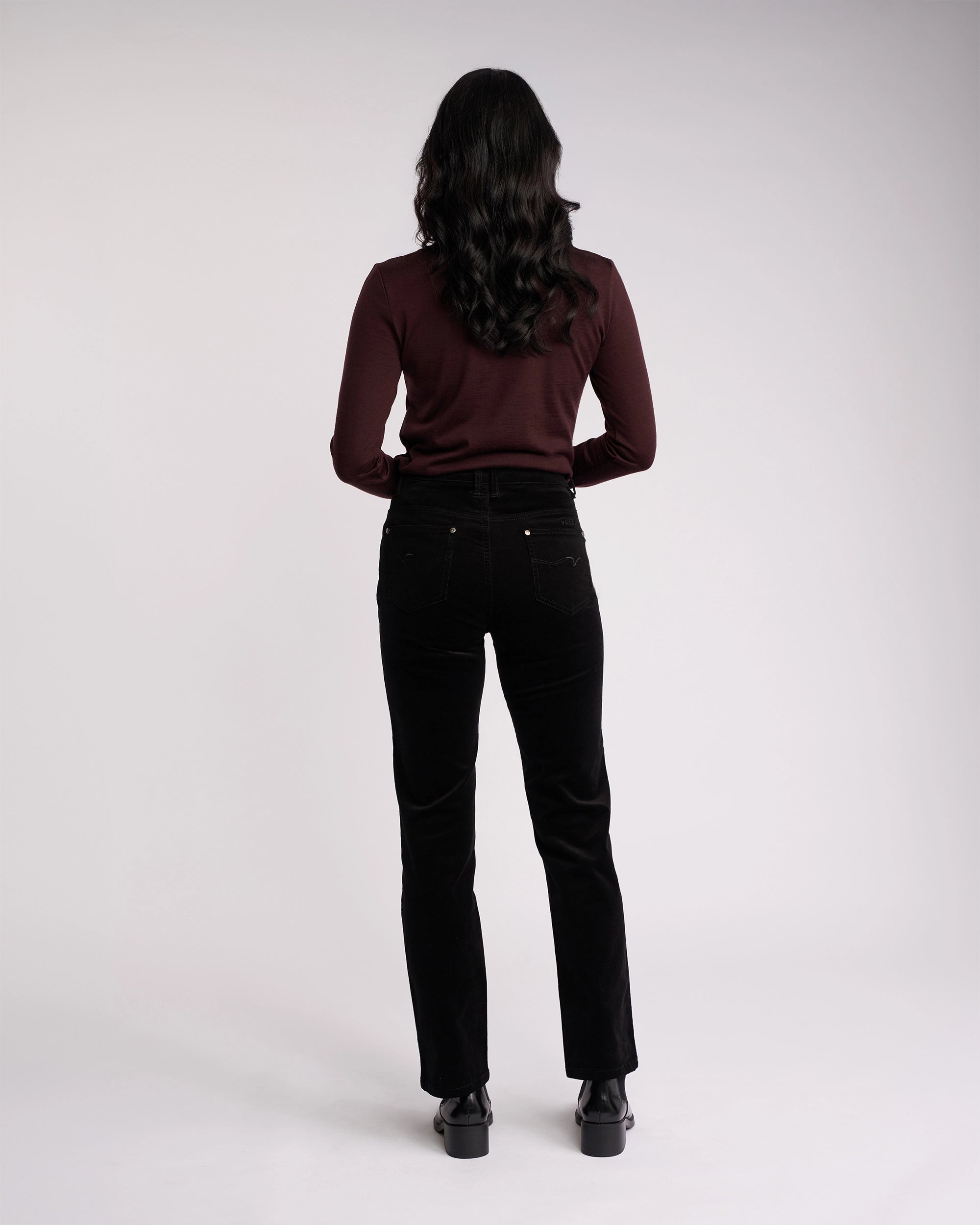 A person with long dark hair, wearing a maroon long-sleeve top and Vassalli Arch Cord Pant in Black, stands facing away against a plain light background—an easy winter style.