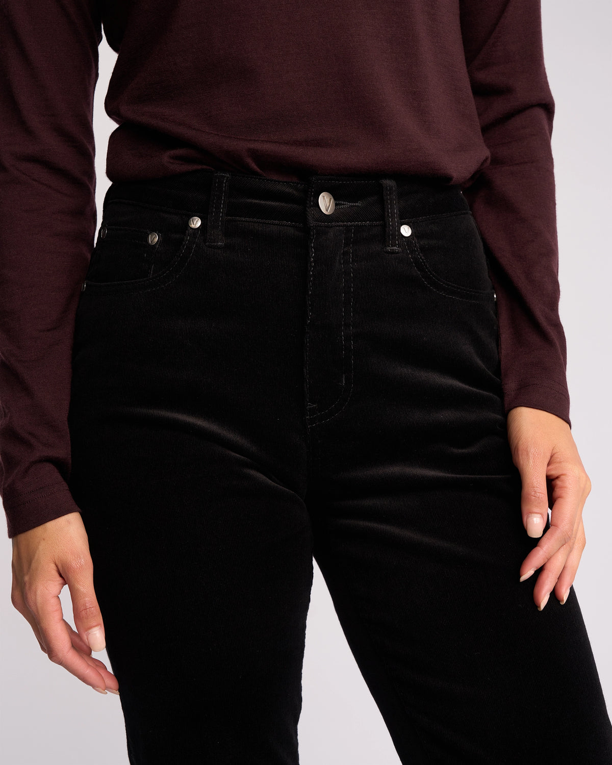 Model shown from shoulders to thighs wearing Vassalli Arch Cord Pant in Black, styled with a long-sleeve dark maroon top; one hand rests by their side, the other on their hip—an ideal winter wardrobe look.