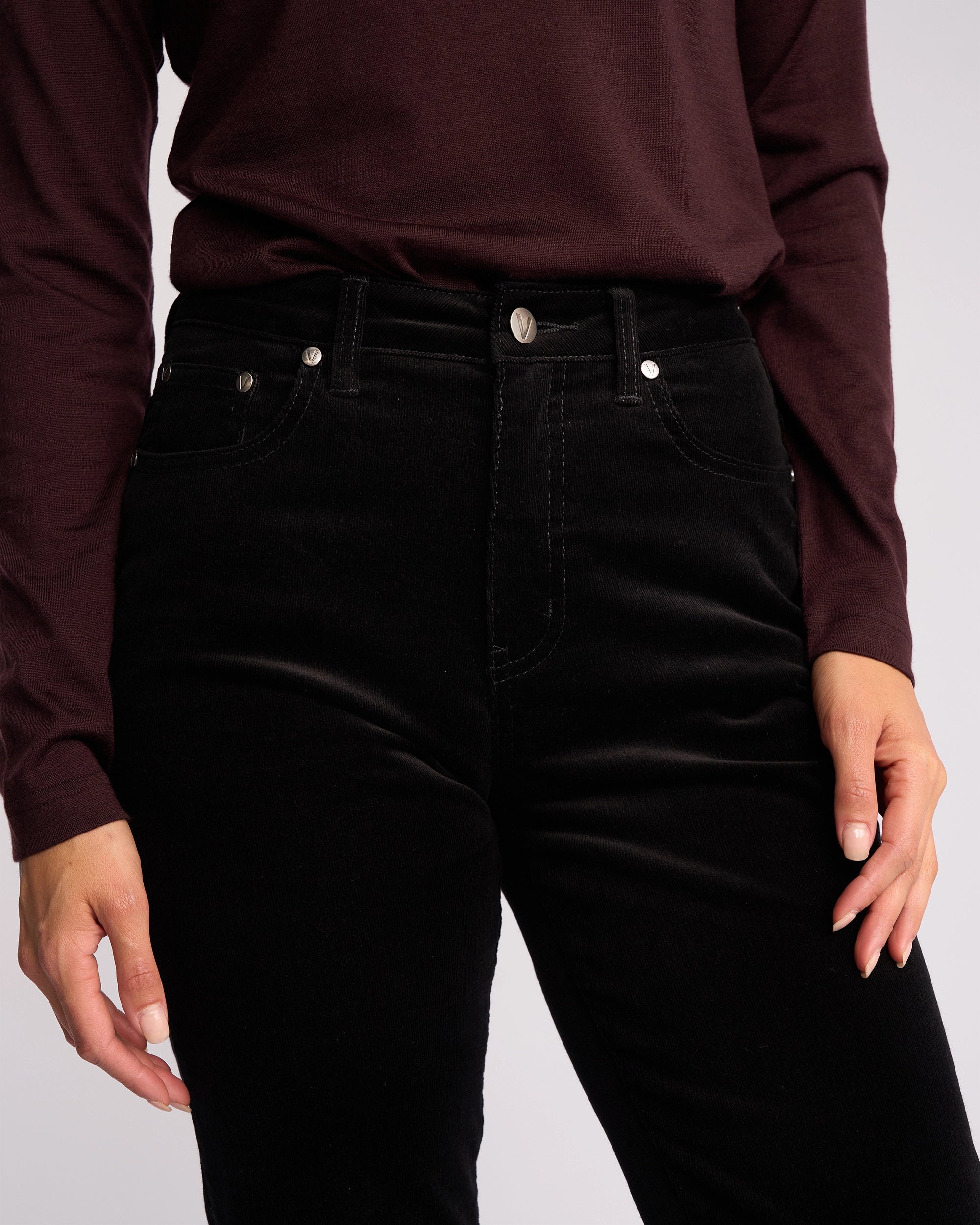 Model shown from shoulders to thighs wearing Vassalli Arch Cord Pant in Black, styled with a long-sleeve dark maroon top; one hand rests by their side, the other on their hip—an ideal winter wardrobe look.