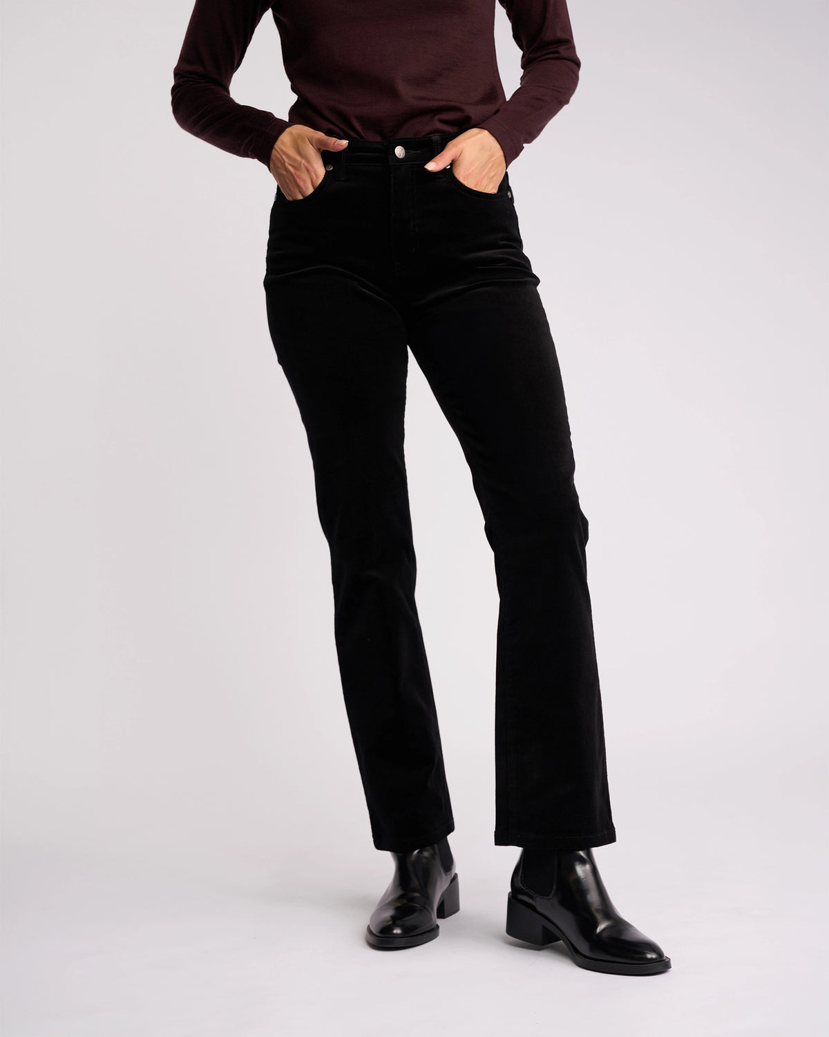 A person models the Vassalli Arch Cord Pant in black pinwale cord, paired with a maroon long-sleeve top and black ankle boots, against a plain light background. The look is chic and winter-ready; the person's head is not shown in the image.