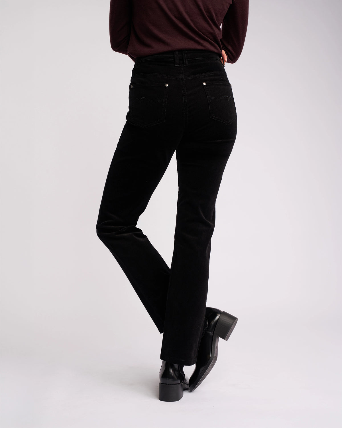 A person wearing Vassalli Arch Cord Pant - Black in pinwale cord, a long-sleeve burgundy top, and black heeled boots stands with legs crossed, facing away from the camera against a plain white background—ideal for winter.