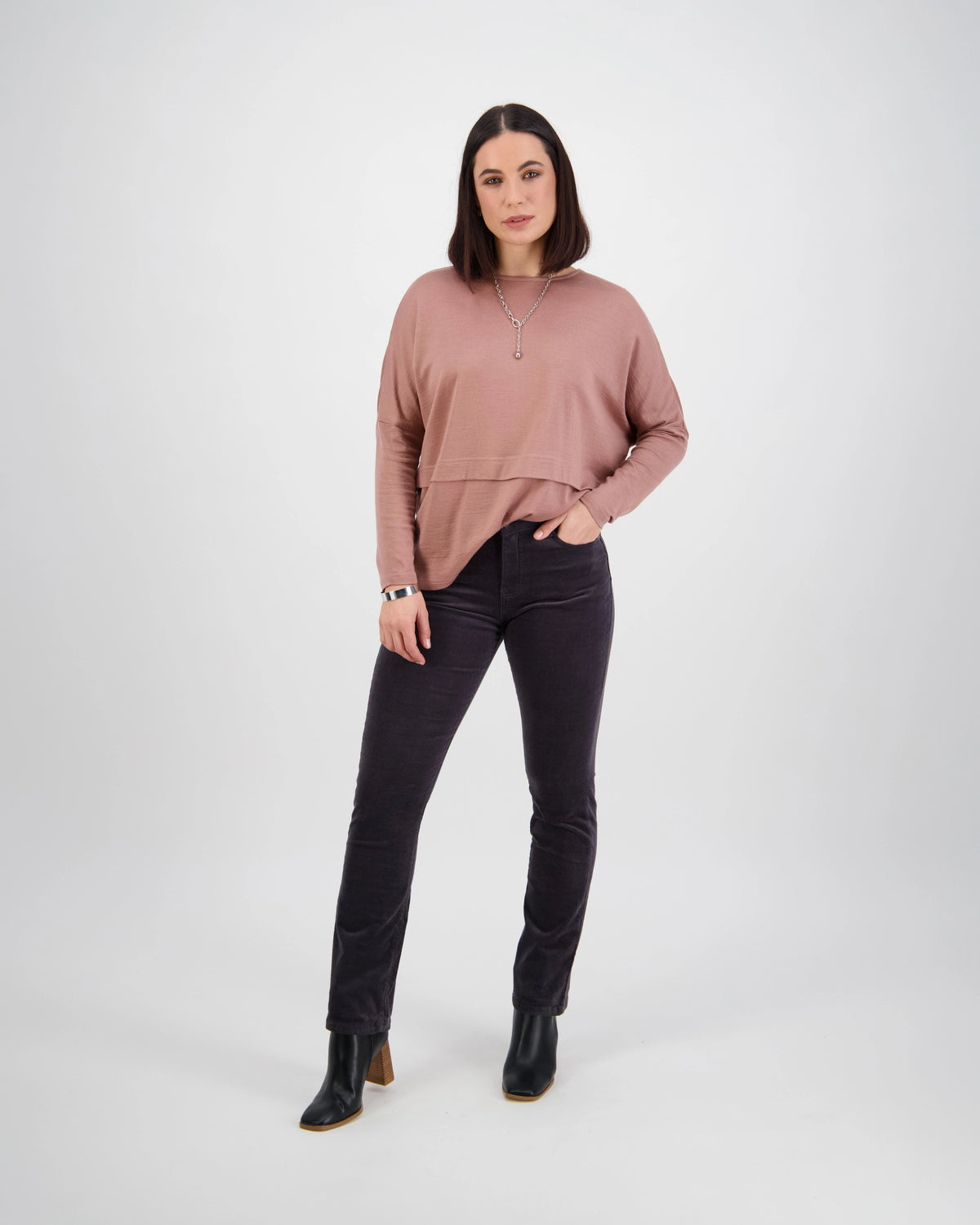 A woman with straight brown hair poses against a white background wearing the Vassalli Arch Cord Pant in Charcoal with a dusty pink long-sleeve top, black ankle boots, and a necklace—an ideal look to refresh your winter wardrobe.