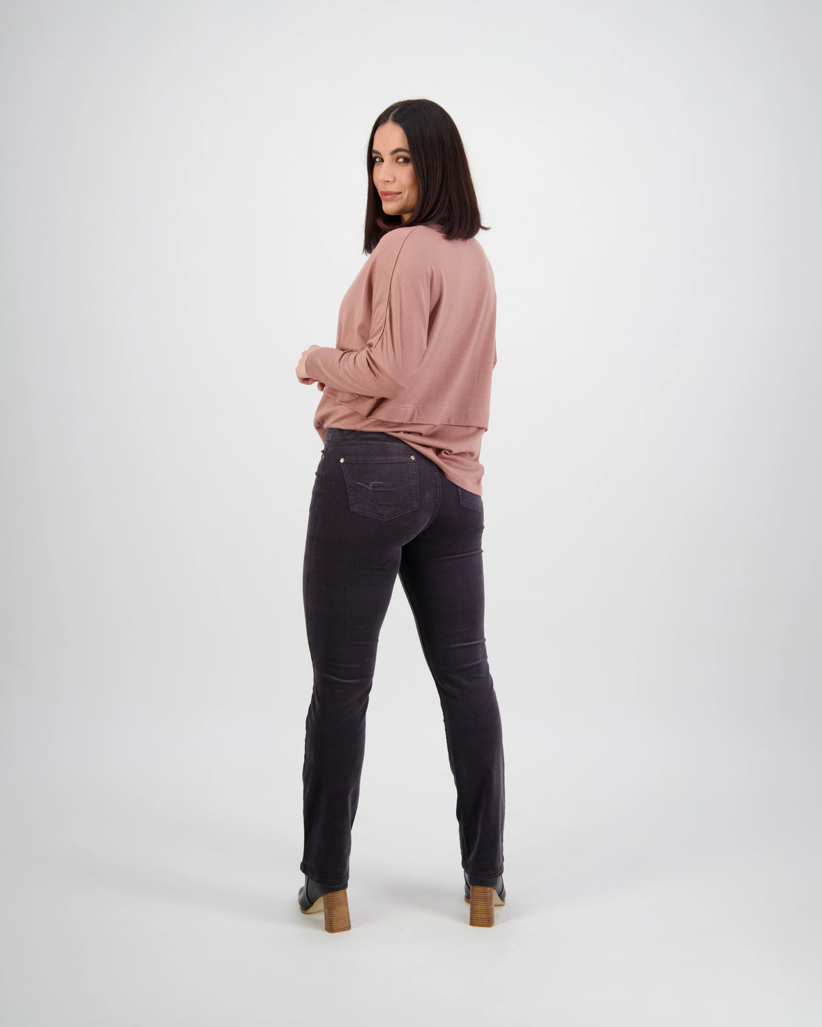 A woman with shoulder-length dark hair stands facing backward, looking over her shoulder. She wears a mauve long-sleeve top and Vassalli Arch Cord Pant in Charcoal, paired with heeled shoes against a plain white background.