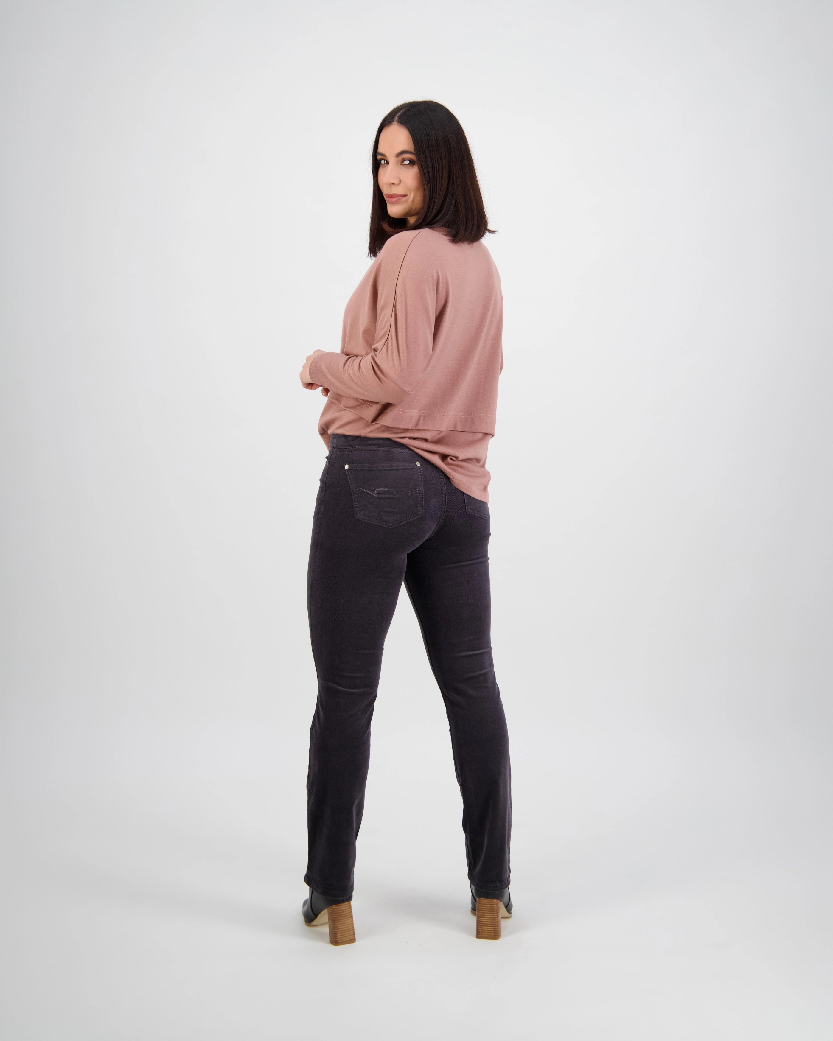 A woman with shoulder-length dark hair stands facing backward, looking over her shoulder. She wears a mauve long-sleeve top and Vassalli Arch Cord Pant in Charcoal, paired with heeled shoes against a plain white background.