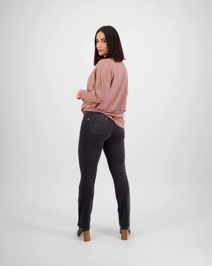 A woman with shoulder-length dark hair stands facing backward, looking over her shoulder. She wears a mauve long-sleeve top and Vassalli Arch Cord Pant in Charcoal, paired with heeled shoes against a plain white background.