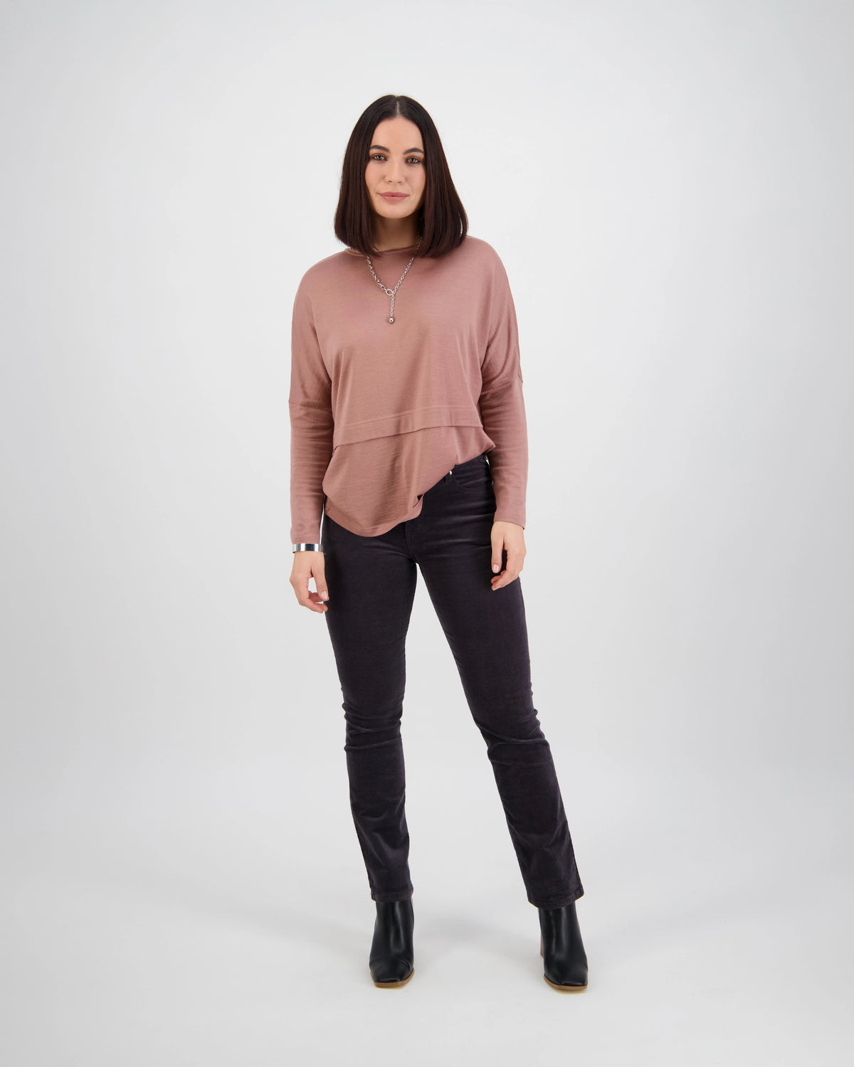 A woman with straight, shoulder-length brown hair stands against a white background, wearing Vassalli’s Arch Cord Pant in Charcoal—a chic addition to any winter wardrobe.