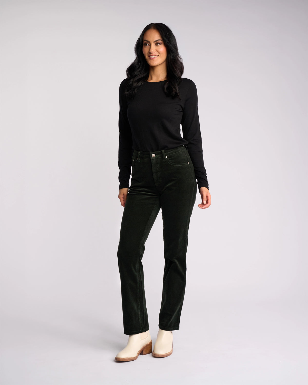 A woman with long dark hair, dressed in a black long-sleeve top, Vassalli Arch Cord Pant - Forest, and ivory ankle boots, stands against a light background, smiling and glancing to the side.