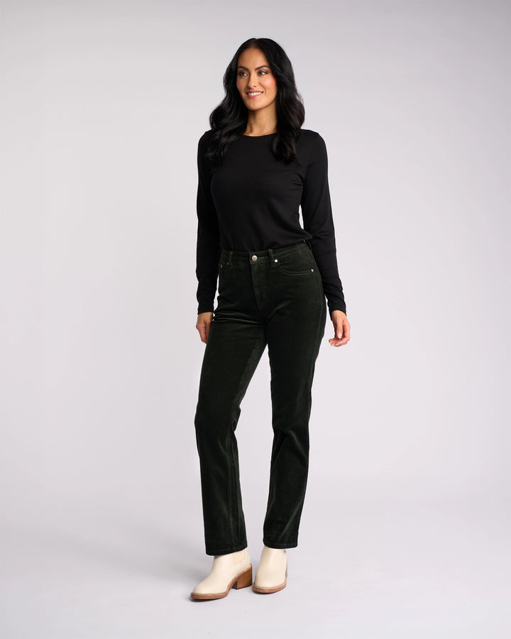 A woman with long dark hair, dressed in a black long-sleeve top, Vassalli Arch Cord Pant - Forest, and ivory ankle boots, stands against a light background, smiling and glancing to the side.