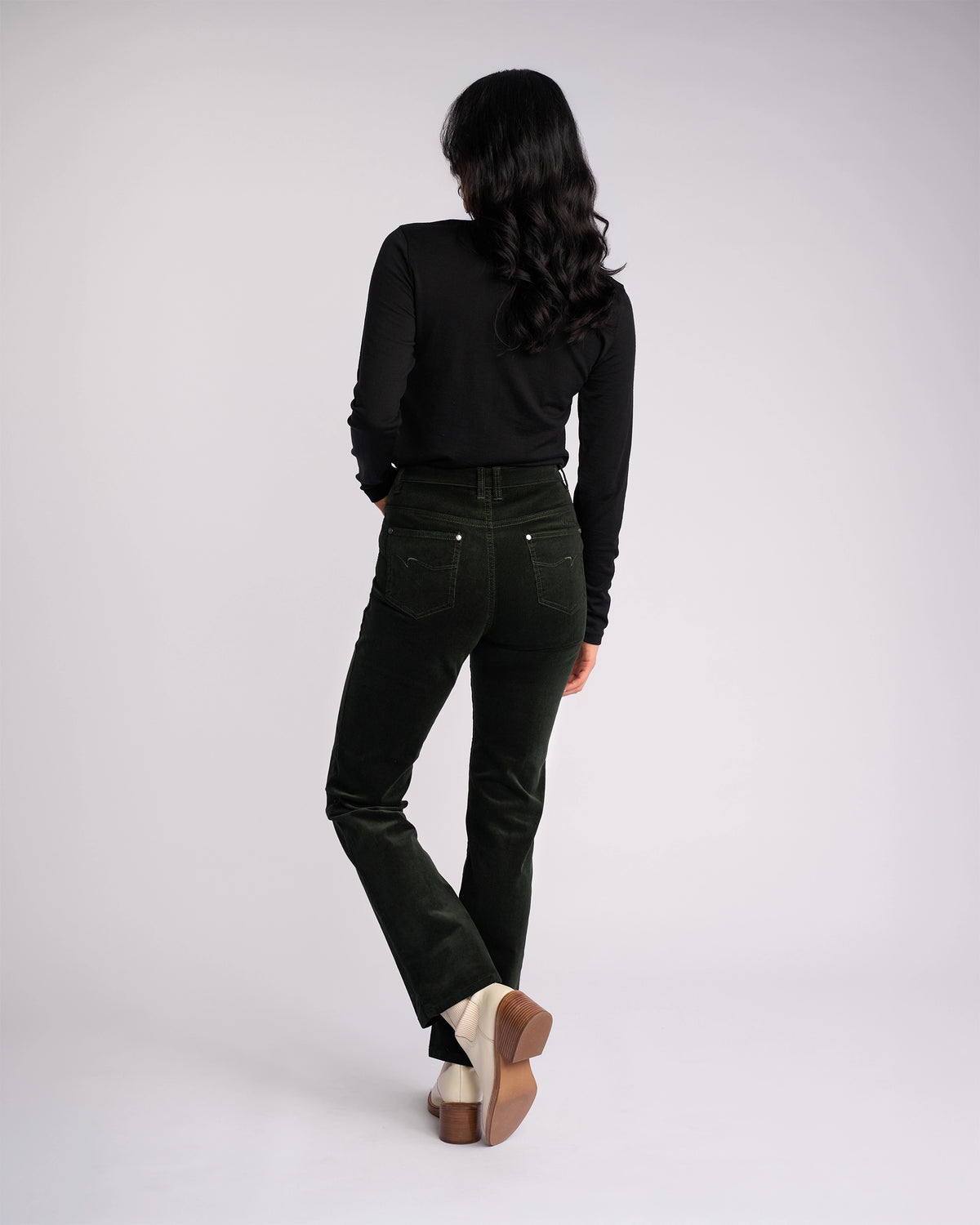 A woman with long wavy dark hair wears a black long-sleeve top, Vassalli Arch Cord Pant - Forest, and light-colored heels as she stands facing away in a studio with a plain light background—ideal for your winter wardrobe.