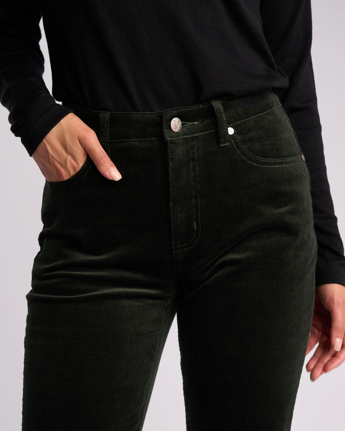 A person wearing a black long-sleeve shirt and Vassalli’s Arch Cord Pant in Forest stands with one hand in their pocket against a plain background, highlighting this stylish winter essential.