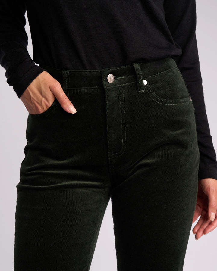 A person wearing a black long-sleeve shirt and Vassalli’s Arch Cord Pant in Forest stands with one hand in their pocket against a plain background, highlighting this stylish winter essential.