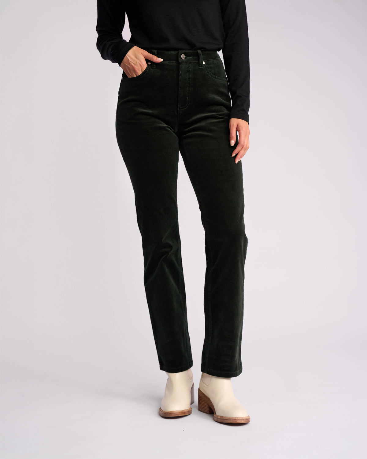 A model wears the Vassalli Arch Cord Pant in Forest, styled with a black long-sleeve top and light ankle boots, posing against a plain light background. The model’s head is not visible in the image.