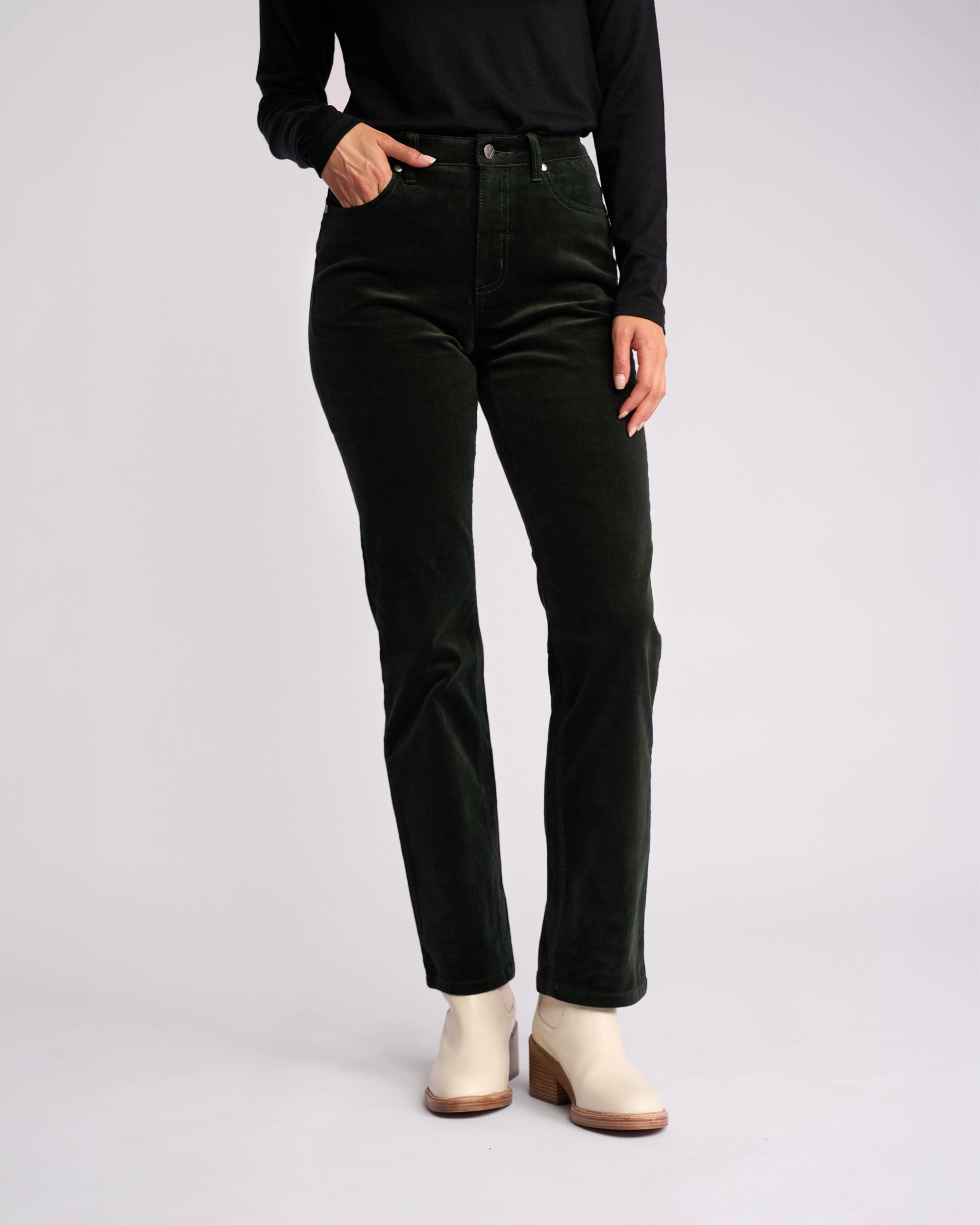 A model wears the Vassalli Arch Cord Pant in Forest, styled with a black long-sleeve top and light ankle boots, posing against a plain light background. The model’s head is not visible in the image.