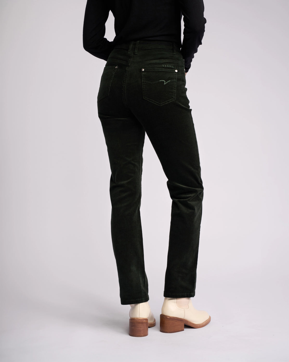 A person in Vassalli’s Arch Cord Pant - Forest stands with their back to the camera, styled with a black long-sleeve top and light beige ankle boots—an ideal look for a winter wardrobe against a plain background.