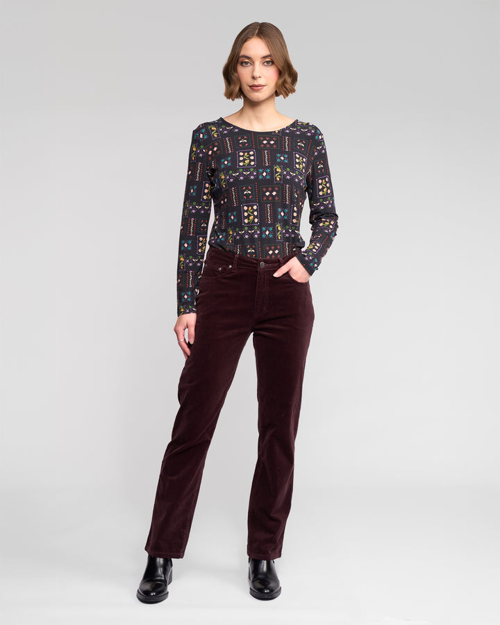 A woman stands against a plain background wearing the Vassalli Arch Cord Pant in Merlot, paired with a long-sleeved patterned top and black ankle boots, one hand in her pocket and a neutral expression.