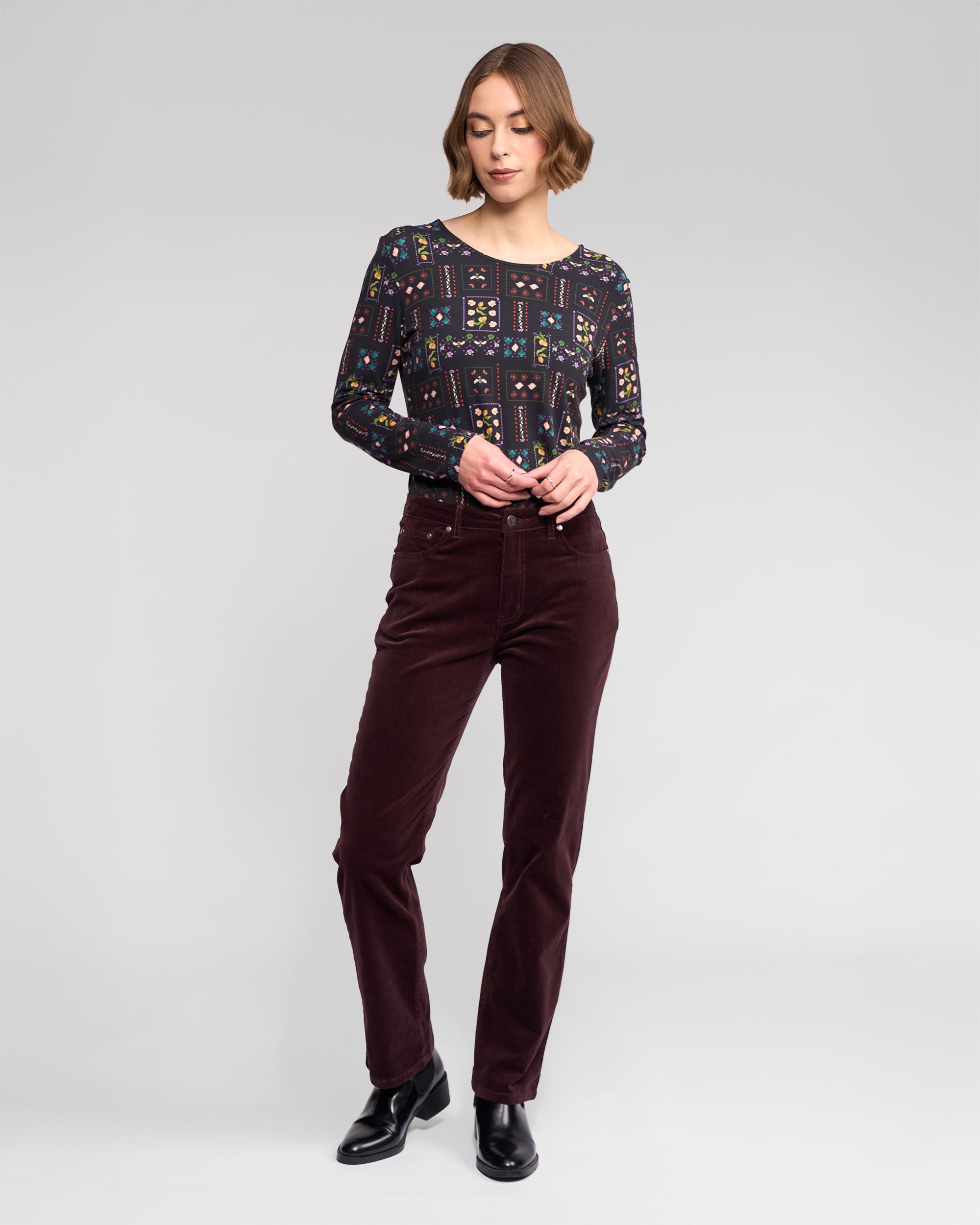 A woman stands against a plain light gray background, wearing a patterned long-sleeve top, black ankle boots, and Vassalli Arch Cord Pant in Merlot. She looks down with her hands near her waist.