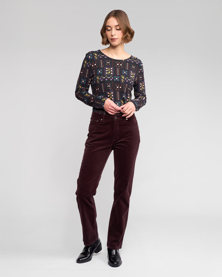A woman stands against a plain light gray background, wearing a patterned long-sleeve top, black ankle boots, and Vassalli Arch Cord Pant in Merlot. She looks down with her hands near her waist.