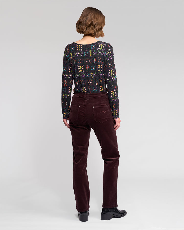 A person with short brown hair is seen from behind, wearing a patterned long-sleeve top, black shoes, and Vassalli’s Arch Cord Pant in Merlot, standing against a plain light gray background.