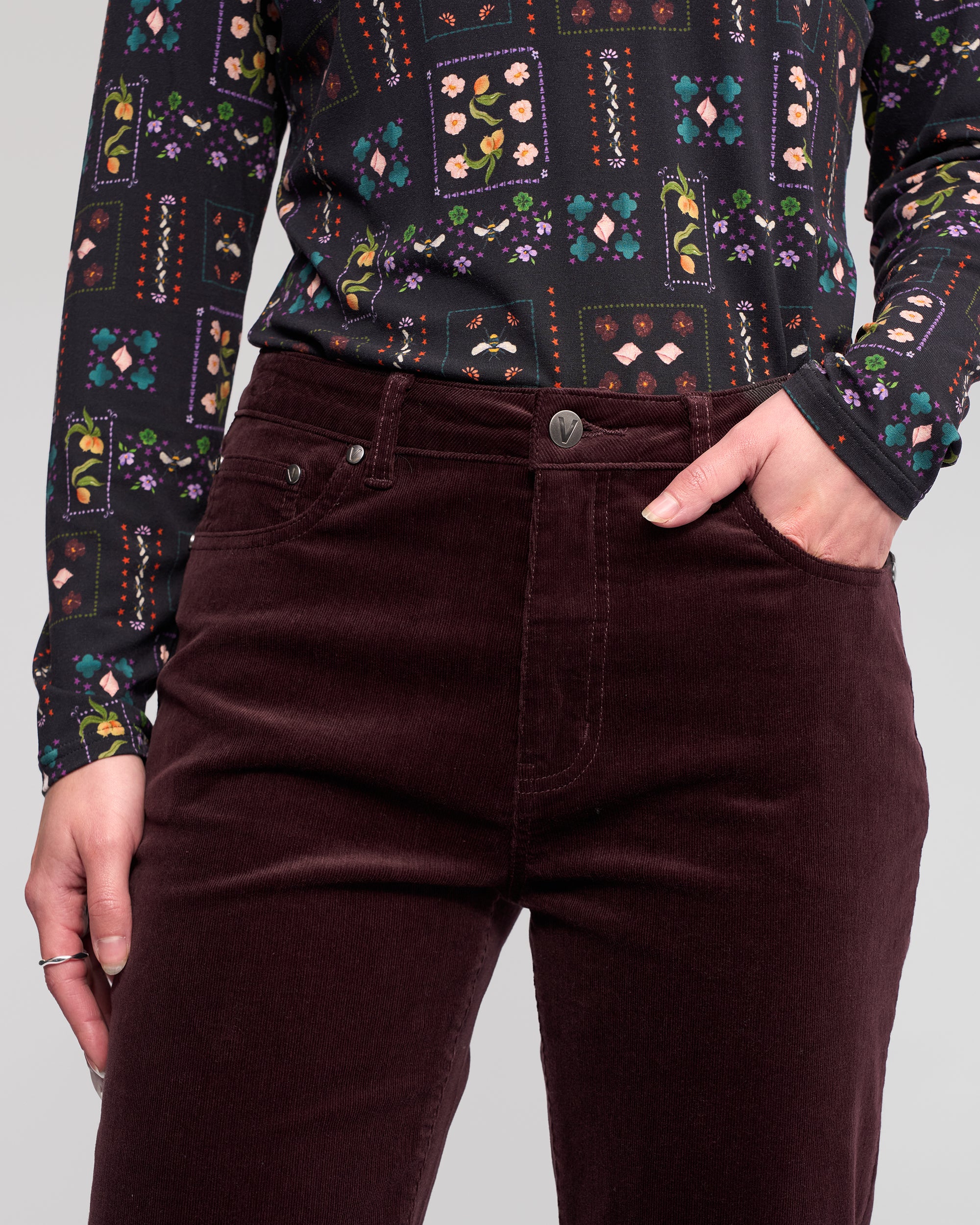A person wearing Vassalli’s Arch Cord Pant in Merlot and a long-sleeve black top with a colorful geometric and floral pattern stands with one hand in their pocket.