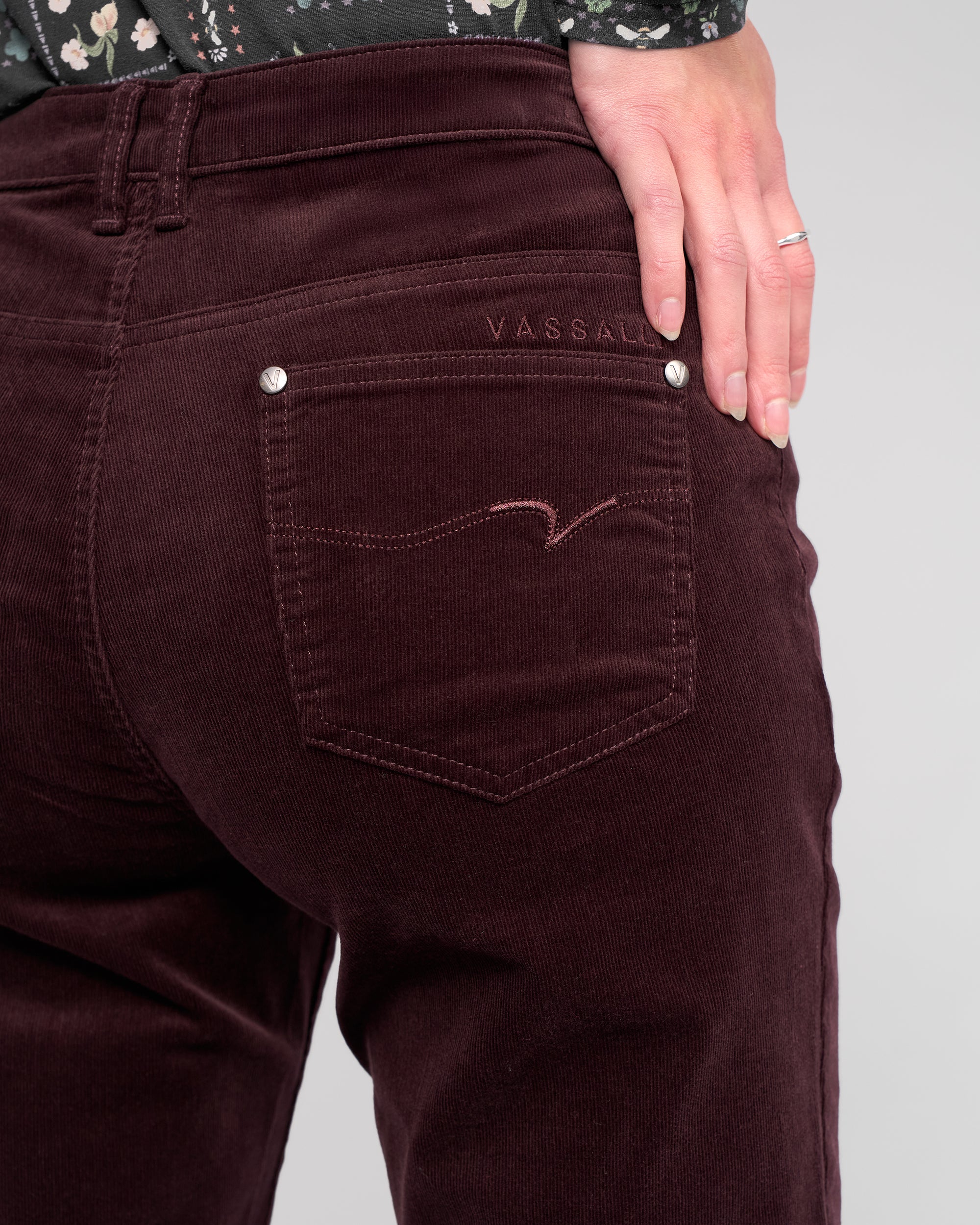 A close-up of a person wearing Vassalli's Arch Cord Pant in Merlot, featuring straight-leg corduroy and VASSALLI embroidery above the back pocket. One hand with a silver ring rests on their hip against a plain light background.