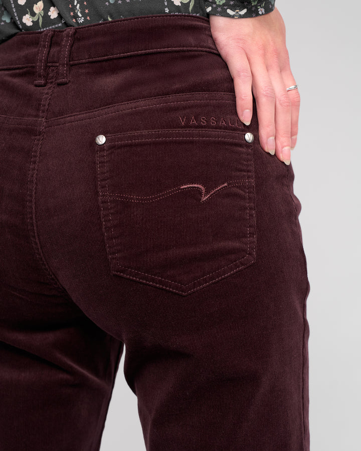 A close-up of a person wearing Vassalli's Arch Cord Pant in Merlot, featuring straight-leg corduroy and VASSALLI embroidery above the back pocket. One hand with a silver ring rests on their hip against a plain light background.