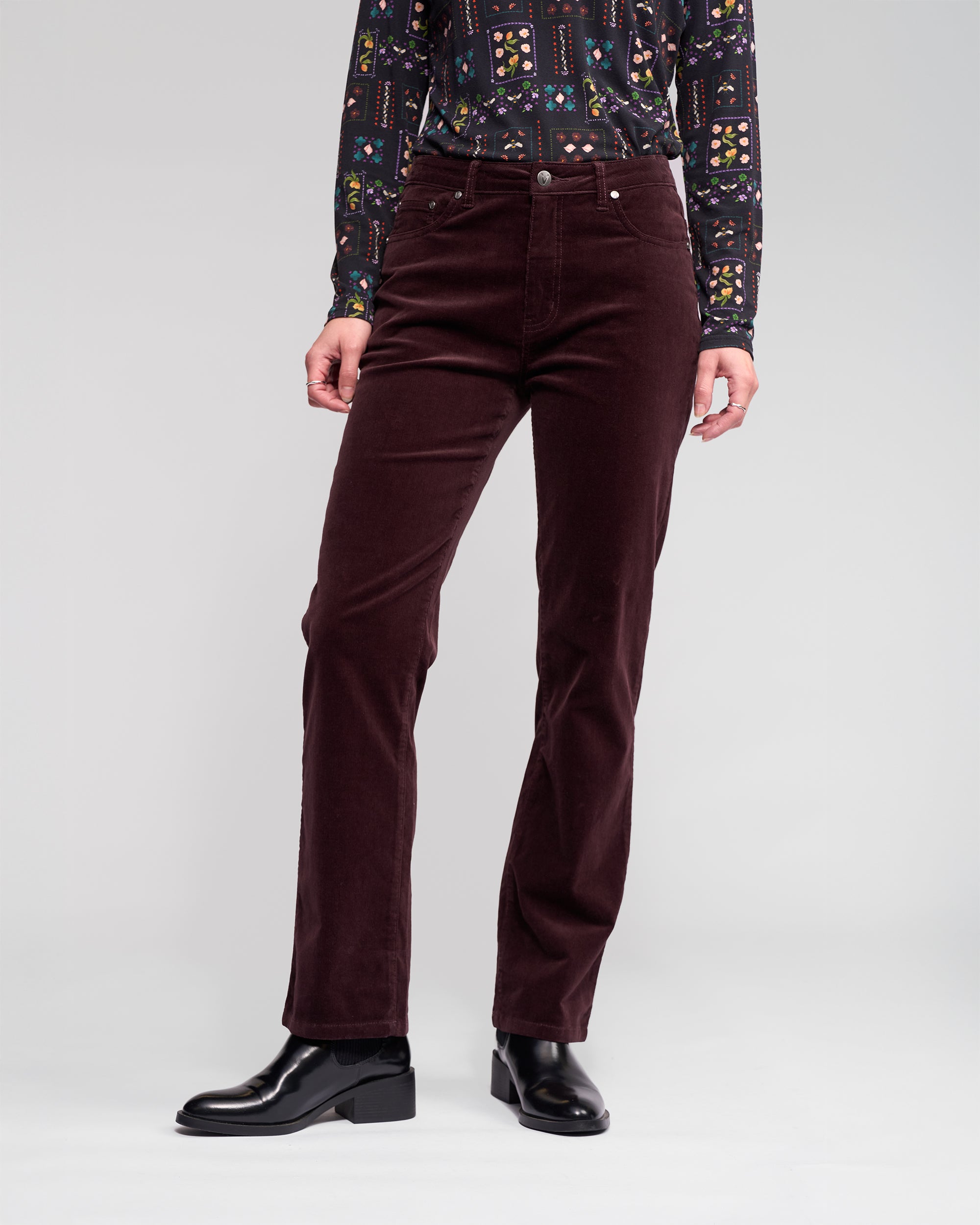 A person wearing a patterned long-sleeve shirt, Vassalli Arch Cord Pant in Merlot, and black ankle boots stands against a plain light gray background.