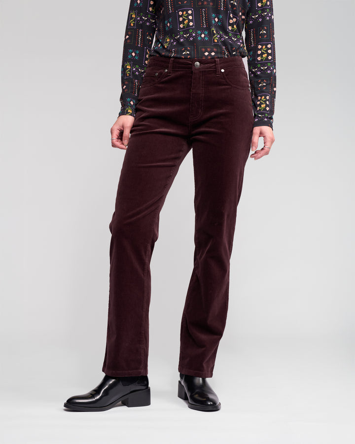A person wearing a patterned long-sleeve shirt, Vassalli Arch Cord Pant in Merlot, and black ankle boots stands against a plain light gray background.