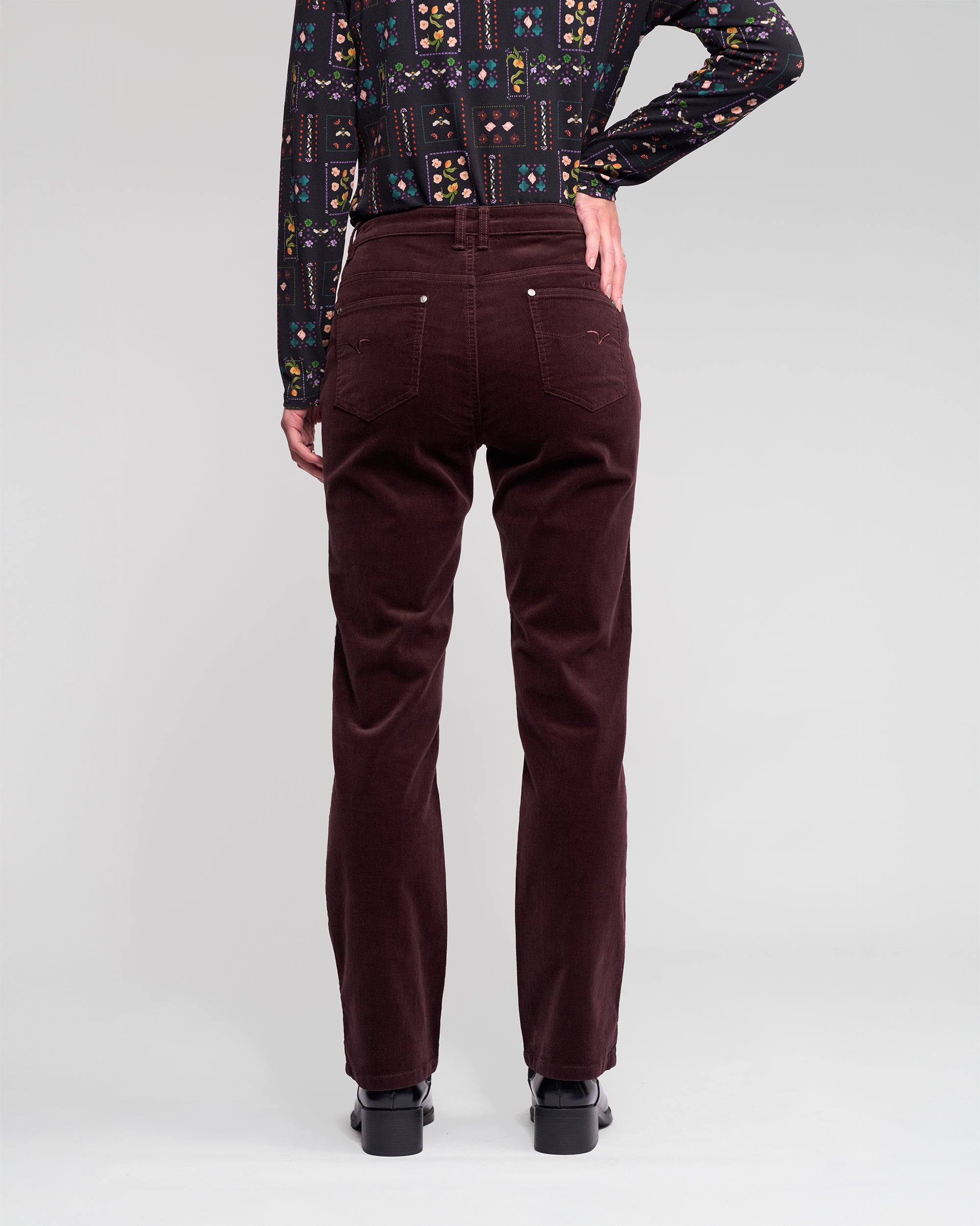 A person stands with their back to the camera, wearing Vassalli’s Arch Cord Pant in Merlot, black heeled shoes, and a long-sleeve shirt featuring colorful geometric patterns.