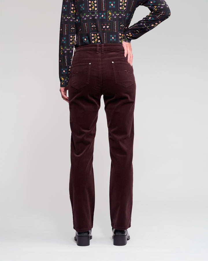 A person stands with their back to the camera, wearing Vassalli’s Arch Cord Pant in Merlot, black heeled shoes, and a long-sleeve shirt featuring colorful geometric patterns.
