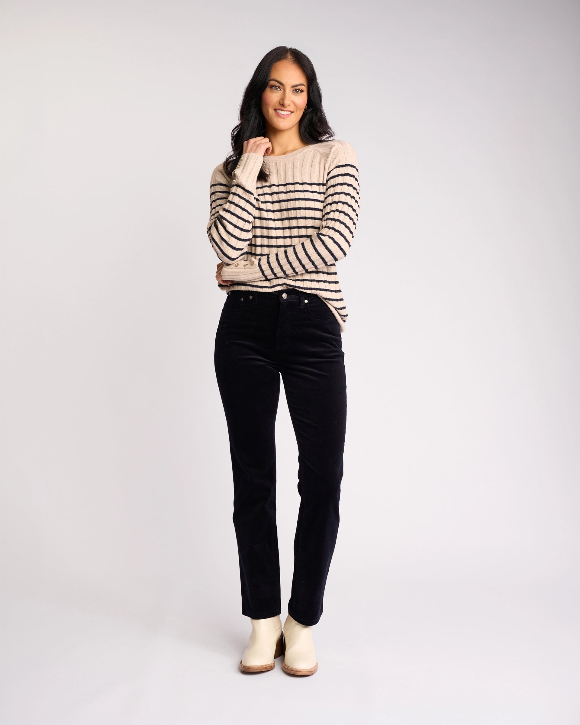 A woman with long dark hair smiles, wearing a beige sweater with black stripes, Vassalli's Arch Cord Pant in Midnight, and light-colored shoes—a stylish winter outfit against a plain, light background.