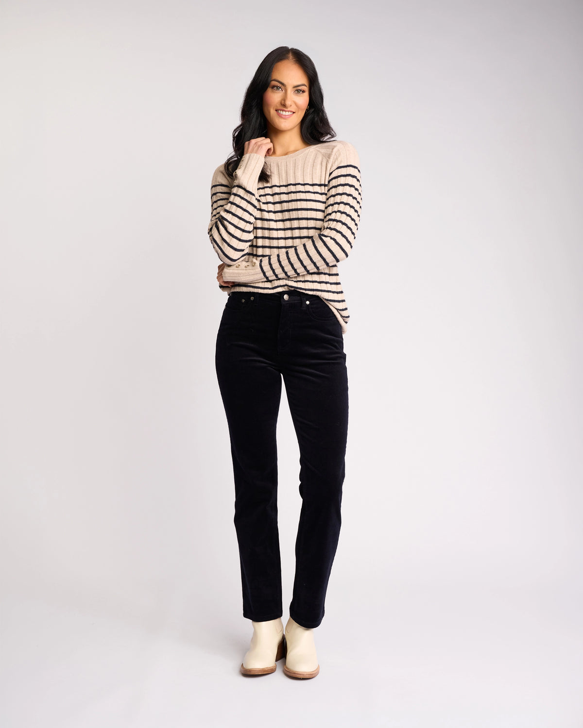 A woman with long dark hair smiles, wearing a beige sweater with black stripes, Vassalli's Arch Cord Pant in Midnight, and light-colored shoes—a stylish winter outfit against a plain, light background.