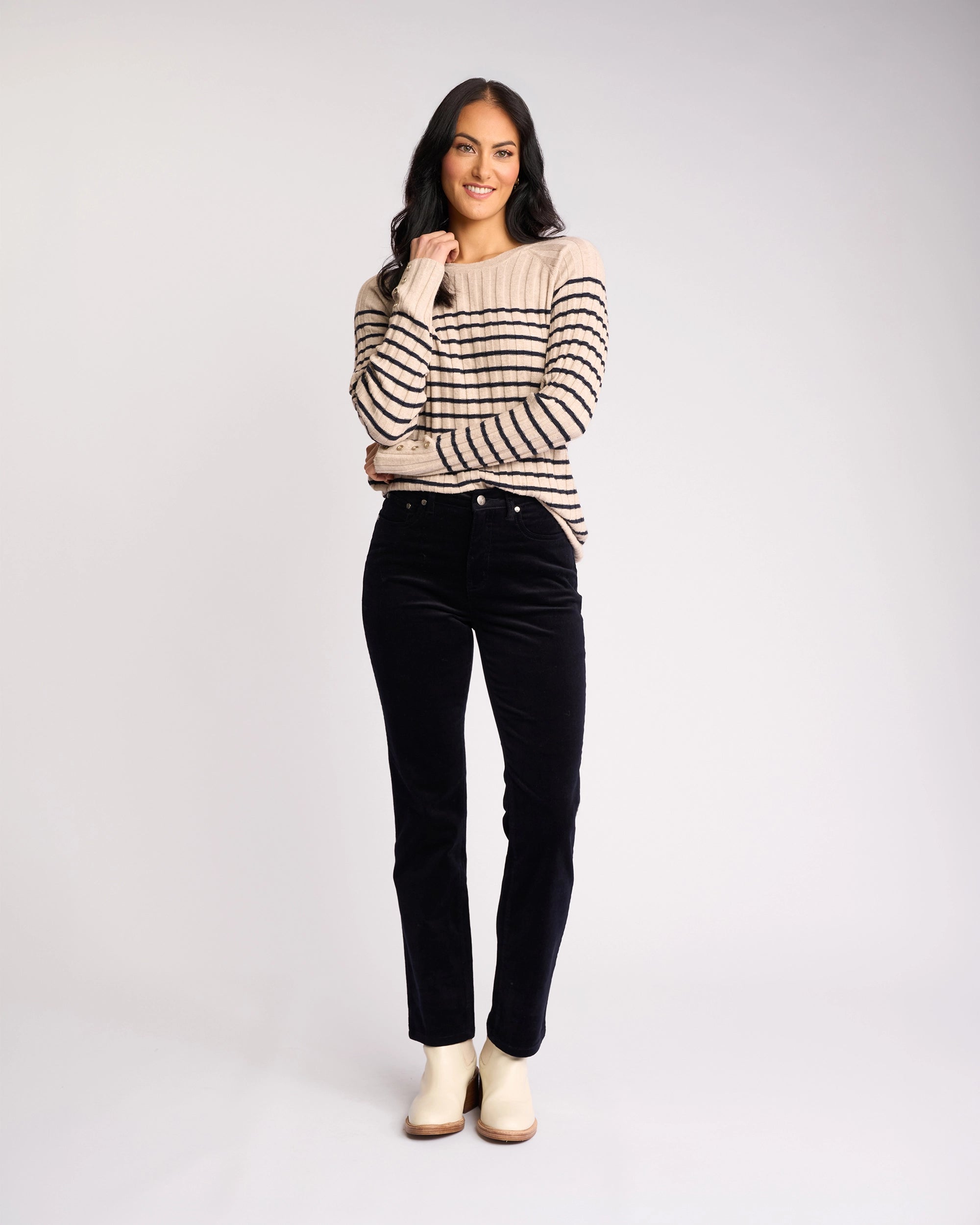 A woman with long dark hair smiles, wearing a beige sweater with black stripes, Vassalli's Arch Cord Pant in Midnight, and light-colored shoes—a stylish winter outfit against a plain, light background.