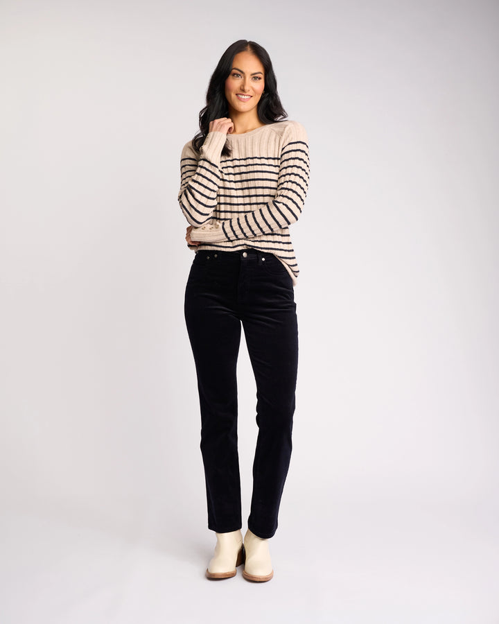 A woman with long dark hair smiles, wearing a beige sweater with black stripes, Vassalli's Arch Cord Pant in Midnight, and light-colored shoes—a stylish winter outfit against a plain, light background.