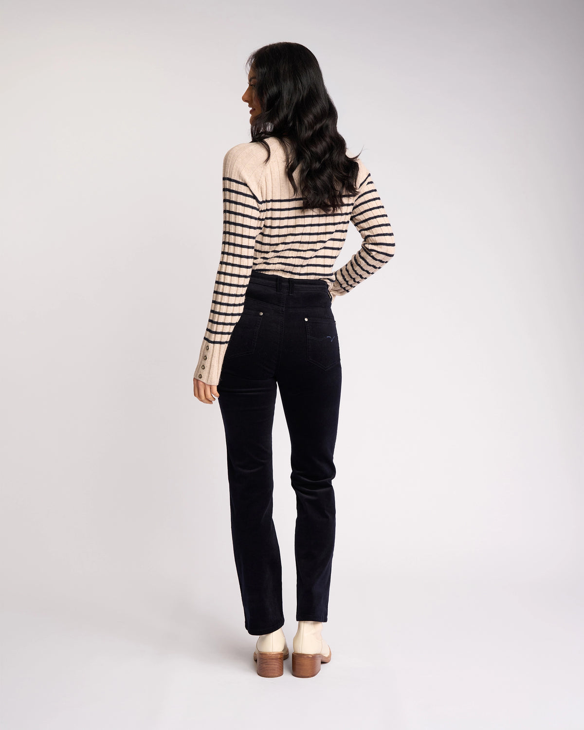 A woman with long dark hair stands facing away, styled in a beige and black striped sweater, Vassalli's Arch Cord Pant - Midnight, and beige heeled shoes—an effortlessly chic look for your winter wardrobe.