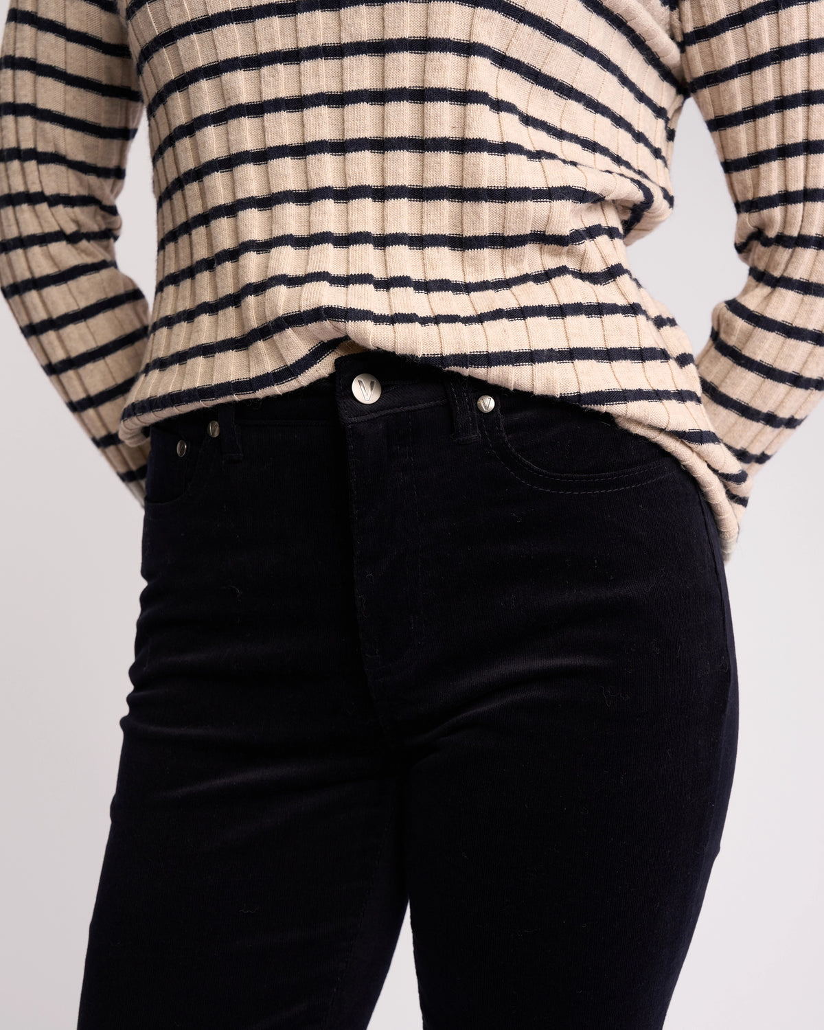 A person wears a cream and black striped long-sleeve shirt with high-waisted black jeans—swap the jeans for Vassalli's Arch Cord Pant - Midnight in soft pinwale cord to add texture to your winter look.