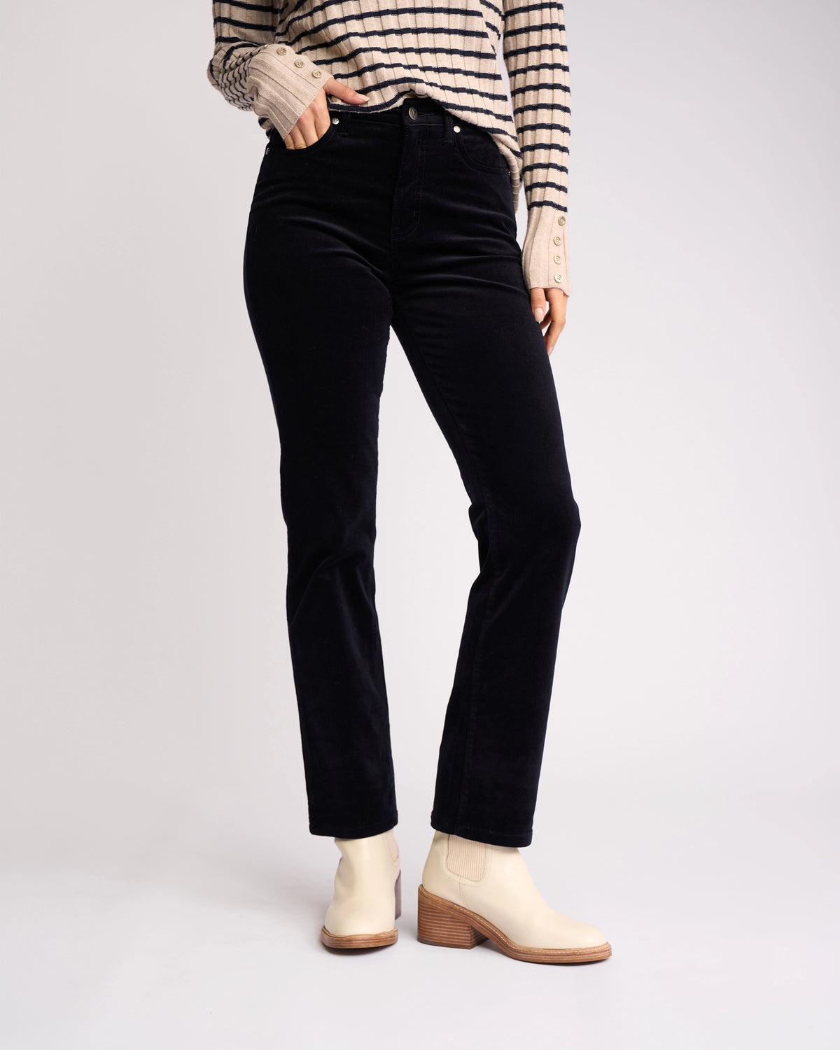 A person models the Vassalli Arch Cord Pant in Midnight—straight-leg black cords—paired with a striped long-sleeve shirt and light ankle boots. Their hand is in their pocket, head out of frame, against a white background. Perfect for winter style.